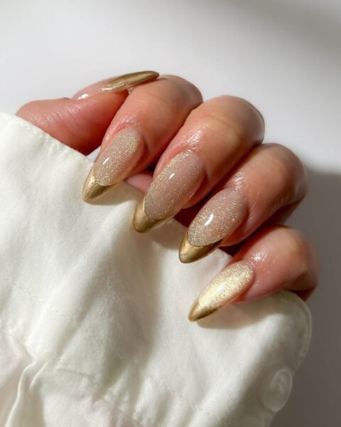 Ekskluzywny i lśniący manicure w kształcie migdała/szpicu. Płytka pokryta jest mleczną/transparentną bazą z delikatnym, iskrzącym brokatem (efekt "Sparkling Nude"), a końcówki paznokci wykończone są metalicznym złotym frenchem. Dłoń modelki w białej, eleganckiej koszuli. Luksusowa inspiracja na manicure wieczorowy lub świąteczny dla kobiet z klasą.