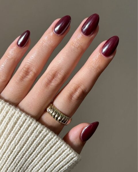 Dłoń modelki z zadbanym manicure. Paznokcie w kształcie migdała/owalu pomalowane modnym, lśniącym lakierem w kolorze głębokiego burgundu z efektem "glazed/glass" (szklanej tafli/glazury). Dłoń otulona jasnym, kremowym swetrem, a na palcu widoczny złoty, prążkowany pierścionek. Klasyczna i luksusowa jesienna inspiracja urodowa.