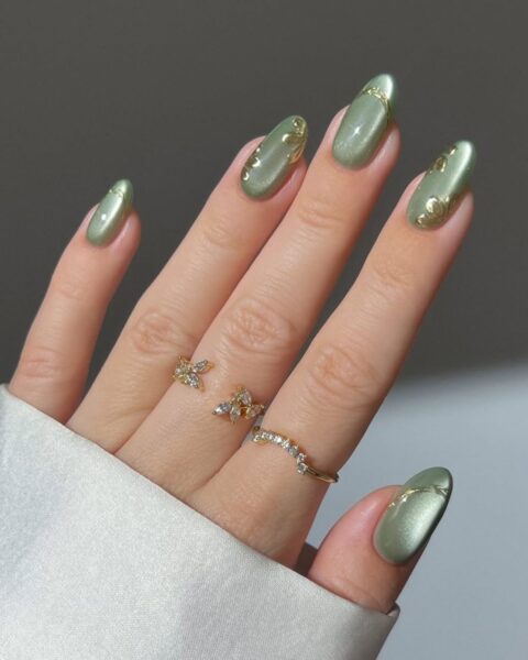 Elegancki manicure w odcieniu szałwiowej zieleni z delikatnym perłowym/satynowym połyskiem. Paznokcie w kształcie owalu/migdała zdobione subtelnymi, złotymi detalami (naklejkami 3D lub ręcznie malowanymi wzorami), imitującymi kwiatowe pędy lub motywy roślinne. Dłoń ozdobiona delikatnymi, złotymi pierścionkami z cyrkoniami (jeden w kształcie motyla), a rękaw białej koszuli dodaje całości klasy.