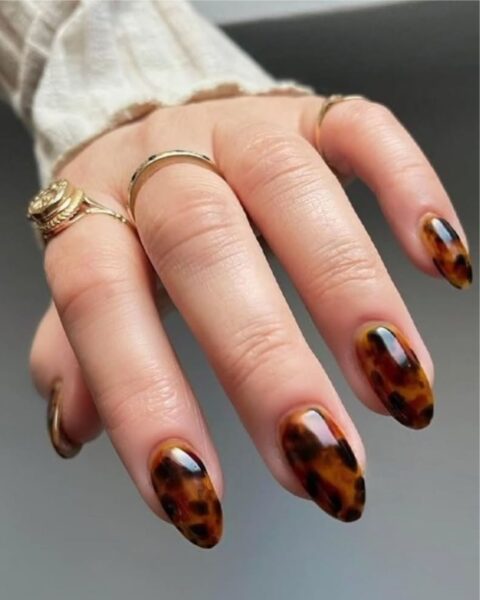 Elegancki, jesienny manicure z modnym wzorem szylkretu (tortoiseshell) w odcieniach ciepłego brązu i czerni. Paznokcie w kształcie owalu/migdała z lśniącym, błyszczącym wykończeniem. Dłoń otulona jasnym, kremowym swetrem z prążkowanej dzianiny, ozdobiona kilkoma złotymi pierścionkami – w tym efektownym, grawerowanym modelem na palcu wskazującym.