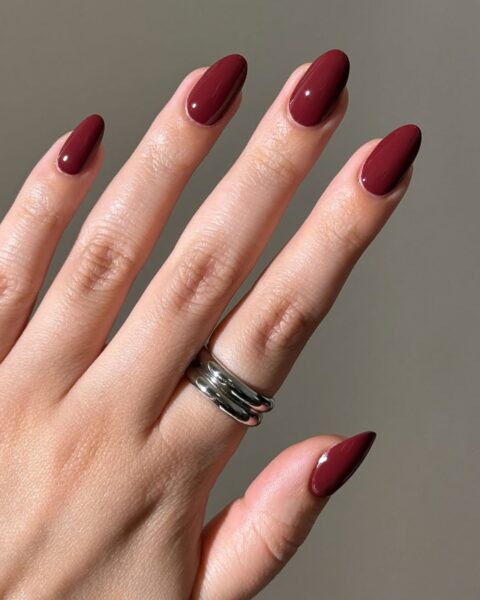 Perfekcyjny manicure w kształcie migdała/szpicu pokryty lśniącym, kryjącym lakierem w głębokim odcieniu klasycznego bordo/wiśniowej czerwieni (burgundu). Kolor jest wyrafinowany i jesienno-zimowy. Na palcu serdecznym widoczny szeroki, srebrny (lub ze stali szlachetnej) pierścionek o nowoczesnym designie. Inspiracja urodowa idealna dla kobiet z klasą.