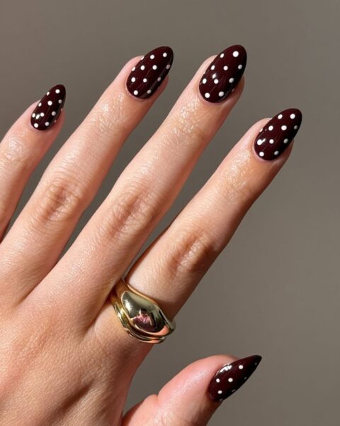 Elegancki manicure z wzorem w kropki (polka dot). Paznokcie w kształcie owalu/migdała pokryte głębokim, lśniącym lakierem w kolorze burgundu lub bardzo ciemnej czekolady, ozdobione równomiernie rozłożonymi, białymi kropkami. Na palcu serdecznym widoczny szeroki, klasyczny złoty pierścień. Ponadczasowa i szykowna inspiracja urodowa.
