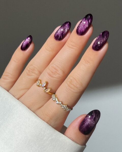 Manicure idealny na jesień i zimę. Paznokcie w kształcie owalu/migdała pomalowane lśniącym lakierem z efektem magnetycznym lub "cat eye" w głębokim, intensywnym odcieniu fioletu lub śliwki, wzbogaconym brokatem/iskrzącym pigmentem. Dłoń ozdobiona delikatnymi złotymi pierścionkami (jeden z kamieniem księżycowym lub cyrkoniami), a fragment białej, eleganckiej koszuli dodaje szyku.