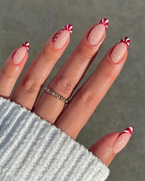 Zbliżenie na dłoń w czerwonym, wełnianym swetrze, prezentującą świąteczny manicure na migdałowych paznokciach. Paznokcie mały i wskazujący są w całości pokryte intensywnym, błyszczącym czerwonym lakierem. Paznokieć środkowy ma naturalną bazę, a jego kontur i serduszko przy nasadzie są obrysowane drobnymi, białymi i czerwonymi kropkami. Paznokieć serdeczny ma również przezroczystą bazę z obrysowanym konturem z biało-czerwonych kropek, nawiązujących do wzoru laski cukrowej.