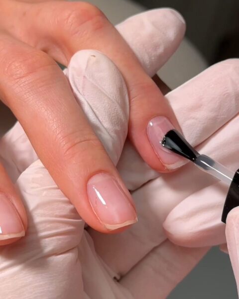 Zbliżenie na dłonie podczas zabiegu pielęgnacyjnego: stylistka nanosi pędzelkiem transparentny preparat – bazę lub odżywkę – na starannie przygotowaną płytkę paznokcia. Zdjęcie emanuje estetyką „clean girl” i pokazuje profesjonalny etap nadawania paznokciom zdrowego blasku.