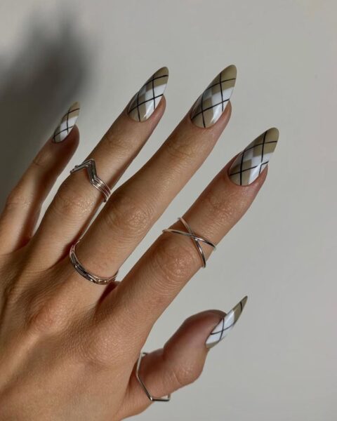 Zbliżenie na dłoń z artystycznym manicure w beżowo-białe geometryczne wzory. Każdy paznokieć ma modny, migdałowy kształt i zdobiony jest motywem argyle (romby) w odcieniach beżu i bieli, przeplatanym czarnymi liniami. Dłoń zdobią minimalistyczne, srebrne pierścionki, w tym jeden w kształcie litery X na palcu serdecznym i jeden geometryczny na kciuku. Tło jest neutralne, jasnoszare.