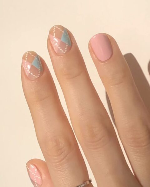 Zbliżenie na zadbaną dłoń z artystycznym manicure na krótkich, naturalnych paznokciach. Płytki palców środkowego i wskazującego ozdobione są geometrycznym wzorem argyle w pastelowych odcieniach różu, błękitu i nude, z delikatnymi, białymi przerywanymi liniami. Paznokieć na palcu serdecznym jest gładki, pomalowany na jednolity pastelowy róż. Na palcu środkowym widać minimalistyczny, wąski, srebrny pierścionek wysadzany drobnymi kryształkami. Dłoń spoczywa na neutralnym, kremowym tle, z widocznym mankietem szarego swetra.