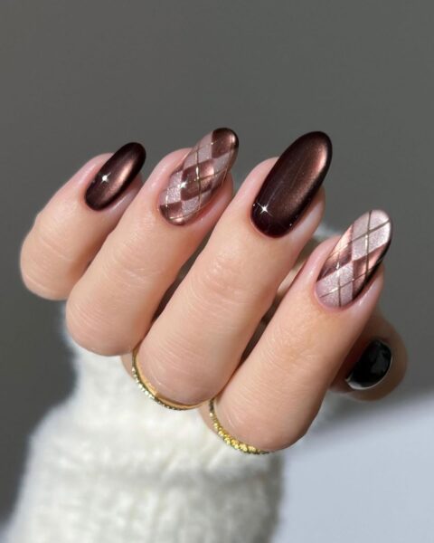 Zbliżenie na dłoń z eleganckim manicure na długich, owalnych paznokciach. Stylizacja łączy głęboki, metaliczny brąz z efektem kociego oka na palcach wskazującym i środkowym, z artystycznym wzorem argyle na pozostałych paznokciach. Motyw rombów na kciuku i palcu serdecznym jest wykonany w odcieniach beżu i metalicznego brązu z cienkimi, złotymi liniami. Na palcach u podstawy widać minimalistyczne, złote pierścionki, a dłoń spoczywa na puszystym, białym swetrze na neutralnym, szarym tle.