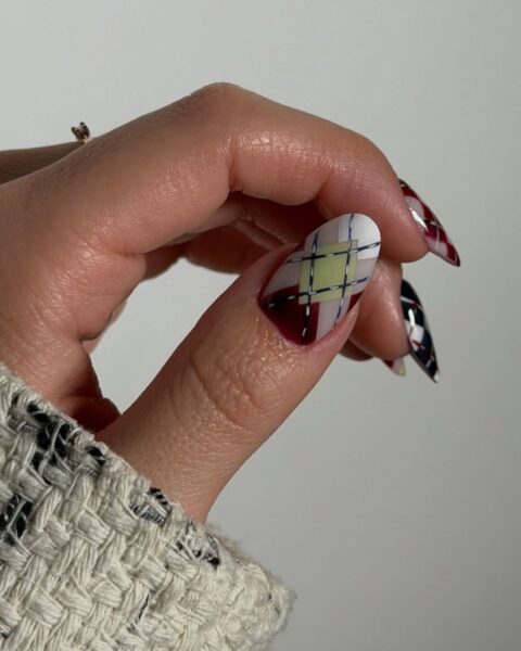 Zbliżenie na dłoń z artystycznym manicure na długich, migdałowych paznokciach. Głównym elementem jest wzór argyle w kolorach burgundu, bieli, błękitu i limonki, który zdobi paznokieć na palcu wskazującym. Pozostałe paznokcie, widoczne w tle, również mają geometryczne wzory. Na palcu widać mały pierścionek, a dłoń jest oparta o puszysty, beżowy sweter. Tło jest neutralne, szare.