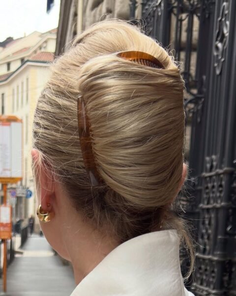 Widok od tyłu na głowę kobiety z jasnymi, blond włosami upiętymi w klasyczny, francuski kok (banał). Fryzura jest zabezpieczona dużą, brązową klamrą (grzebieniem) w stylu szylkretowym. Kobieta ma na sobie białą koszulę i duży, złoty kolczyk w kształcie koła. Tło jest lekko rozmyte (efekt bokeh) i przedstawia fragment europejskiej ulicy z kamienicami oraz ozdobną, czarną, kutą bramą po prawej stronie. Zdjęcie wykonane jest w świetle dziennym.
