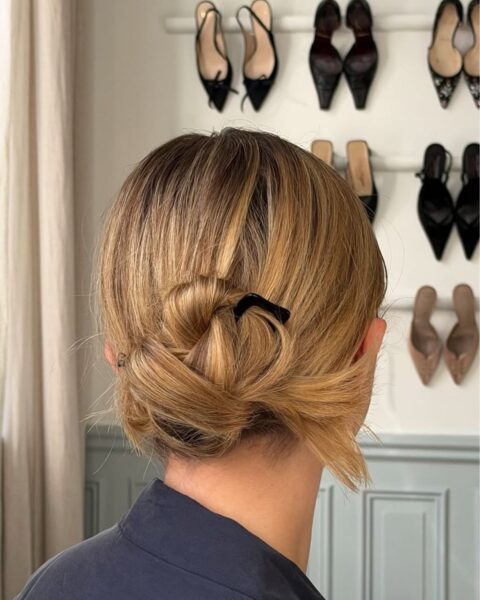 Widok od tyłu na głowę kobiety z jasnymi, miodowo-blond włosami upiętymi w swobodny, teksturowany niski kok typu "messy bun". Fryzura jest spięta małą, czarną klamrą w kształcie serca, widoczną po prawej stronie. Kobieta ma na sobie ciemnoszarą lub granatową bluzkę. Tło stanowi jasne, eleganckie wnętrze z szarą boazerią u dołu i kolekcją damskich butów (czółenka, klapki) wyeksponowanych na półkach na białej ścianie. Po lewej stronie widać fragment jasnej,lnianej zasłony. Zdjęcie jest ostre i dobrze oświetlone.