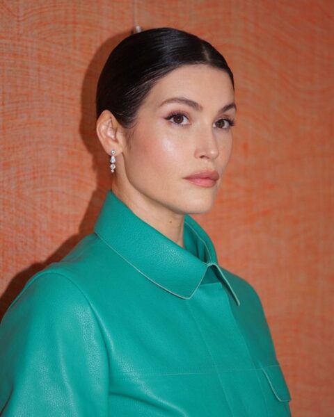 Elegancka kobieta z gładko zaczesanymi do tyłu, ciemnymi włosami upiętymi w niski kok, w zielonym płaszczu i subtelnych kolczykach, na tle ciepłej, pomarańczowej ściany