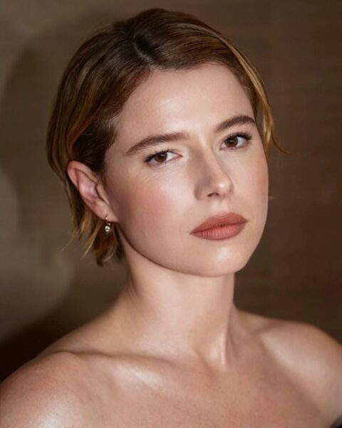 Portret kobiety o jasnej cerze z krótkimi, lekko zaczesanymi na bok włosami w odcieniu jasnego brązu, z naturalnym makijażem i delikatnymi kolczykami, na miękko rozmytym, ciepłym tle.