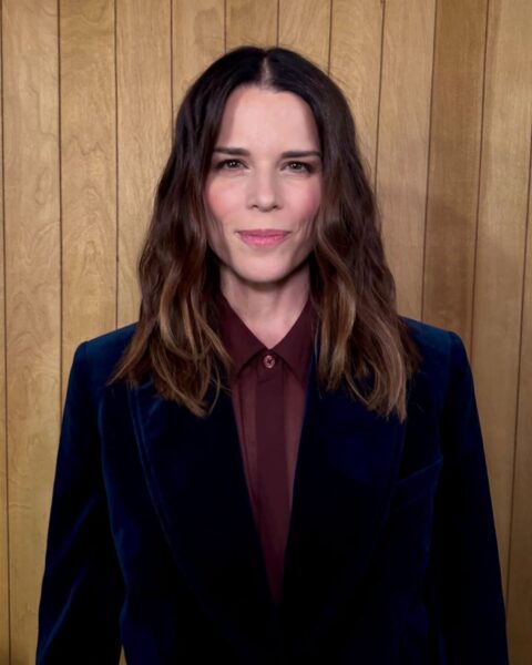 Kobieta o średniej długości, lekko falowanych włosach w odcieniu brązu, ubrana w elegancką granatową marynarkę i bordową koszulę, stojąca na tle drewnianej ściany.