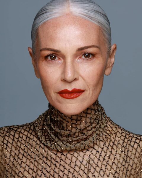 Portret dojrzałej kobiety z krótkimi, gładko zaczesanymi siwymi włosami, wyrazistym czerwonym makijażem ust i subtelnym makijażem oczu, ubranej w elegancką, półtransparentną bluzkę z ozdobną siateczką, na neutralnym tle.