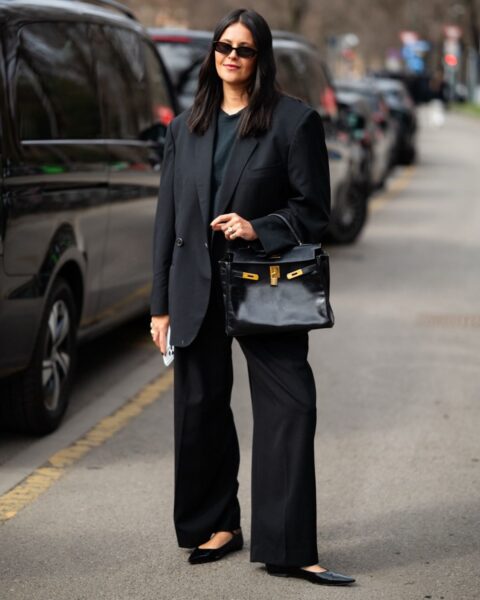 Kobieta stoi na miejskiej ulicy, uśmiechając się lekko do aparatu. Jest ubrana w elegancki, całkowicie czarny strój: dwurzędową marynarkę oversize i dopasowane szerokie spodnie. Pod spodem ma prosty ciemny t-shirt. Nosi ciemne okulary przeciwsłoneczne i subtelną biżuterię. W lewej ręce trzyma luksusową czarną torebkę ze skóry z wyraźnymi złotymi okuciami, w tym charakterystycznym zapięciem i kłódką. W prawej ręce trzyma smartfon. Jej buty to czarne, spiczaste baleriny na płaskiej podeszwie. W tle widać rząd zaparkowanych samochodów na ulicy.