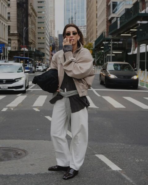 Kobieta w nowoczesnej, warstwowej stylizacji street style na tle miejskiej zabudowy. Ma na sobie beżową kurtkę bomberkę, ciemną koszulę w kratę oraz białe, szerokie spodnie o luźnym kroju. W pasie ma przewiązany szary sweter, a pod pachą trzyma dużą, czarną torebkę o ciekawej teksturze. Całość dopełniają czarne, lakierowane mokasyny oraz wąskie okulary przeciwsłoneczne. Postać stoi na przejściu dla pieszych w otoczeniu wielkomiejskich budynków i samochodów.