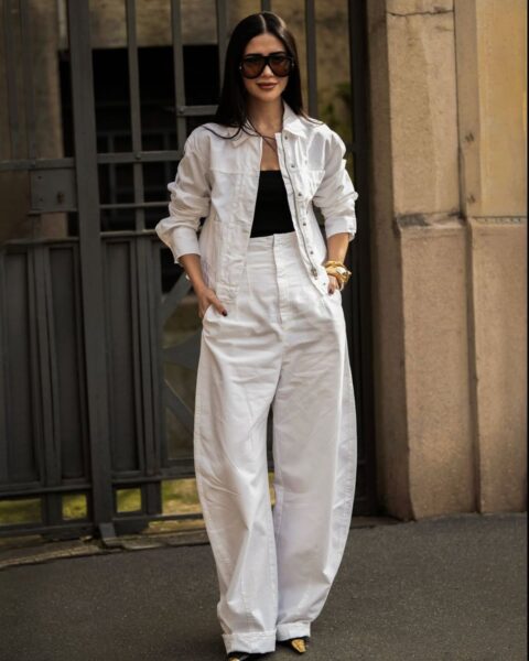 Modna kobieta w białym komplecie składającym się z kurtki i szerokich spodni stoi na ulicy. Pod kurtką ma czarny t-shirt, a na twarzy ciemne okulary przeciwsłoneczne. Jej dłonie spoczywają w kieszeniach spodni. Całość uzupełnia złota biżuteria na nadgarstku i widoczne czarno-złote buty. W tle widać ciemną metalową bramę i fragment budynku.