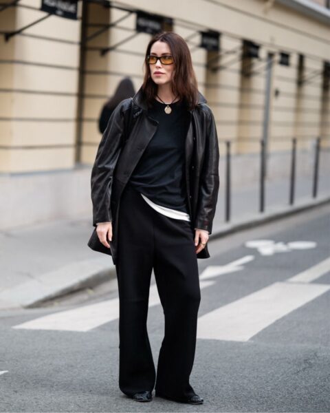 Modna kobieta w czarnej skórzanej kurtce, czarnej bluzce i czarnych szerokich spodniach stoi na ulicy. Na szyi ma naszyjnik z wisiorkiem, a na nosie ciemne okulary przeciwsłoneczne. Tło stanowią kamienice z jasnymi fasadami i czarnymi detalami.