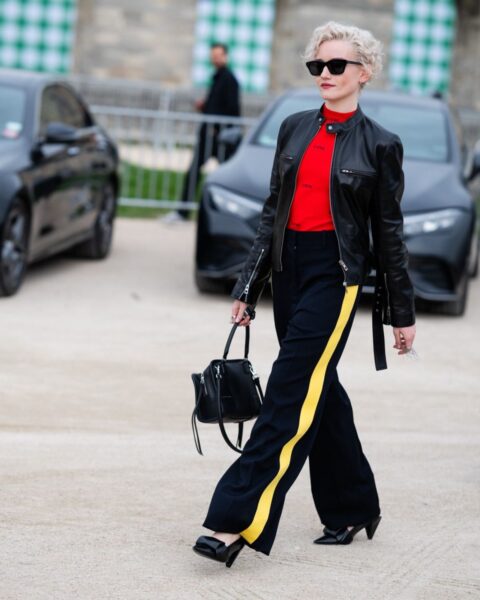 Kobieta o krótkich, jasnych, kręconych włosach idzie pewnym krokiem w nowoczesnej stylizacji. Ma na sobie czarną skórzaną kurtkę motocyklową, dopasowany czerwony golf oraz szerokie, granatowe spodnie z charakterystycznym żółtym lampasem wzdłuż nogawki. Całość uzupełniają czarne okulary przeciwsłoneczne, czarna torebka do ręki oraz czarne czółenka na niewysokim obcasie z ozdobnym elementem z przodu. W tle widać zaparkowane samochody oraz metalowe barierki.