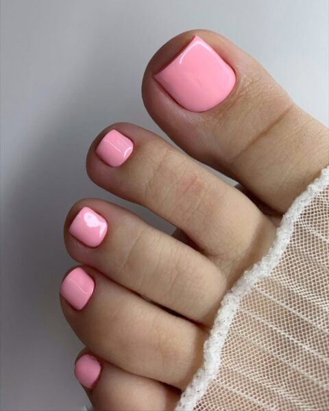 Zdjęcie z bliska przedstawia zadbane kobiece palce u stóp z profesjonalnym pedicure. Paznokcie mają precyzyjny, kwadratowy kształt i są pomalowane jednolitym, intensywnym lakierem w kolorze pudrowego różu o wysokim połysku. Skóra jest gładka, a wokół kostki widoczny jest fragment białej skarpetki wykonanej z siateczki. Całość ujęta na jasnym, neutralnym tle.