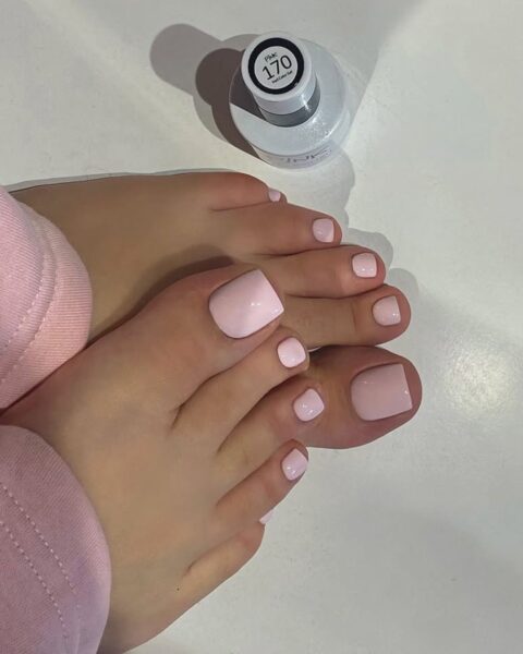 Zdjęcie przedstawia stopy z profesjonalnym pedicure w jasnym, pastelowym odcieniu różu o błyszczącym wykończeniu. Paznokcie mają staranny, kwadratowy kształt. Obok stóp, na białym tle, stoi buteleczka lakieru hybrydowego z numerem 170 na nakrętce. Osoba na zdjęciu ma na sobie jasnoróżowe spodnie dresowe, co tworzy spójną kolorystycznie, estetyczną kompozycję.