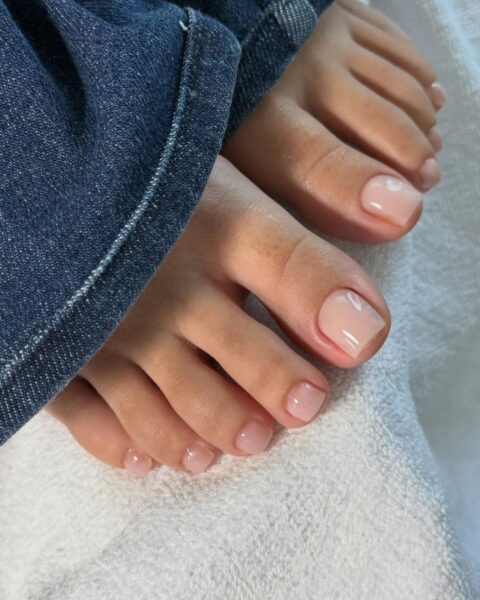 Ujęcie z bliska na stopy z profesjonalnie wykonanym pedicure. Paznokcie mają precyzyjny, kwadratowy kształt i są pokryte lśniącym lakierem hybrydowym w odcieniu mlecznoróżowym typu nude. Stopy spoczywają na białym ręczniku frotte, a obok nich widoczny jest rąbek nogawki niebieskich dżinsów.